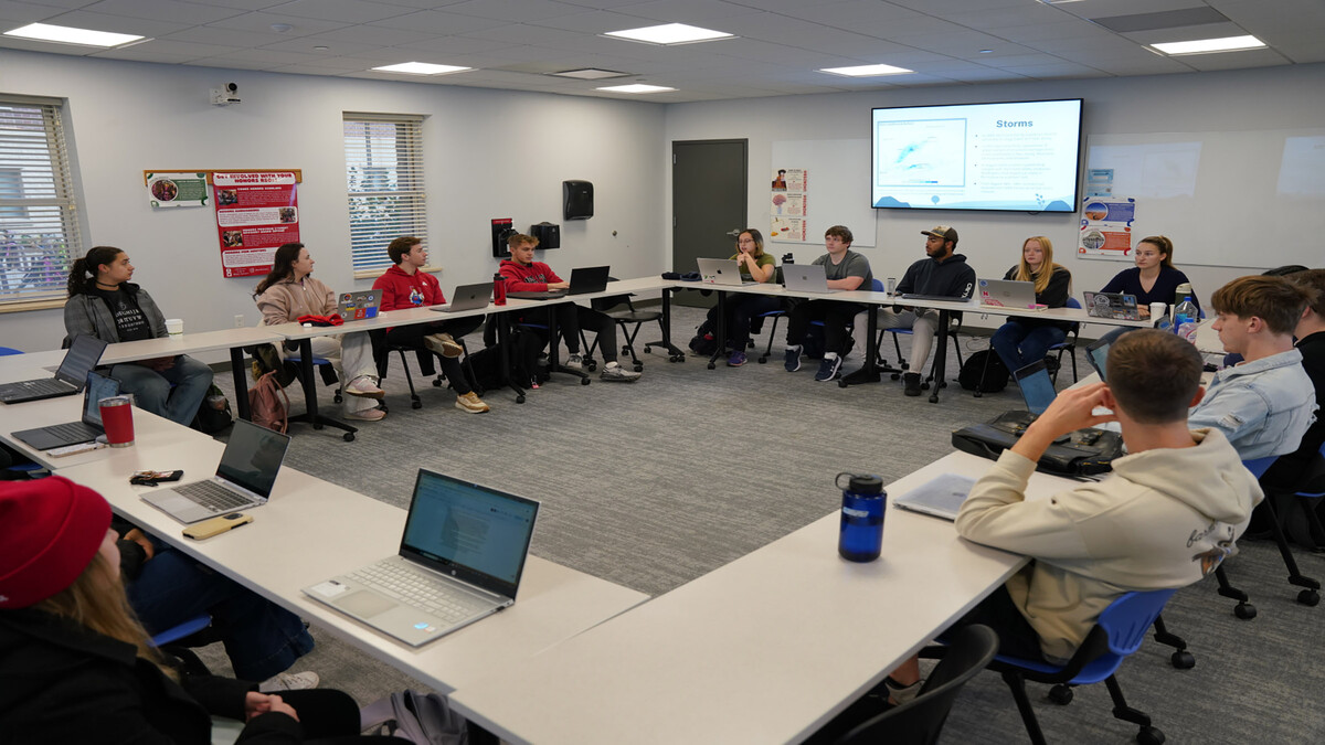 Students sit in a square of tables with their laptops open, in front of a tv screen depicting a slideshow about responding to storms.