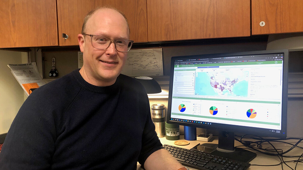 Ian Ratcliffe, a white bald man with glasses, stands in a black long-sleeved t-shirt next to a computer picturing a map he created.