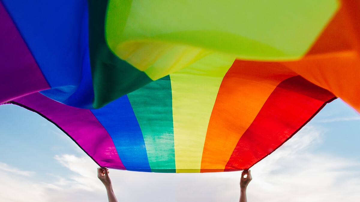 Person holds a pride flag up to flutter in the breeze with the sky in the background.