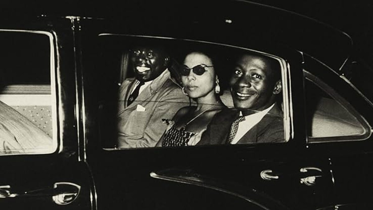 Andreé Blouin sits in the middle of the back seat of a car between two other Black men in suits, in a black and white image.