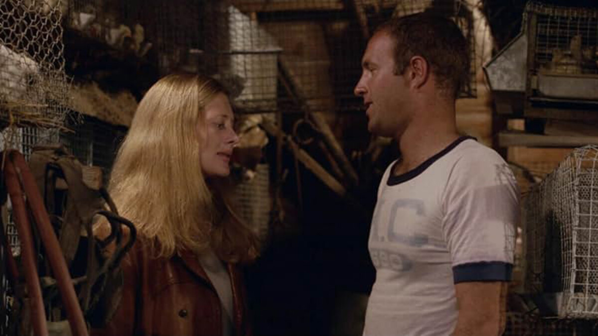 A white blonde woman stands to the left, in front of a taller brunette man in a white t-shirt.