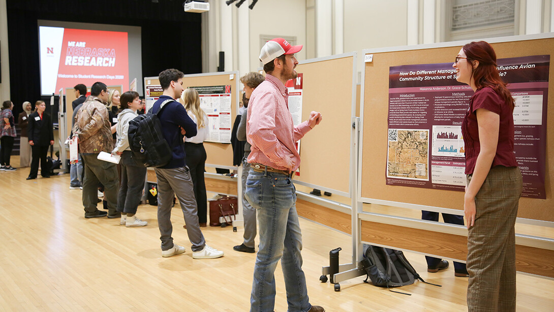 A young man speaks with a student researcher, while the researcher stands next to a research poster in Nebraska Union's Ballroom.