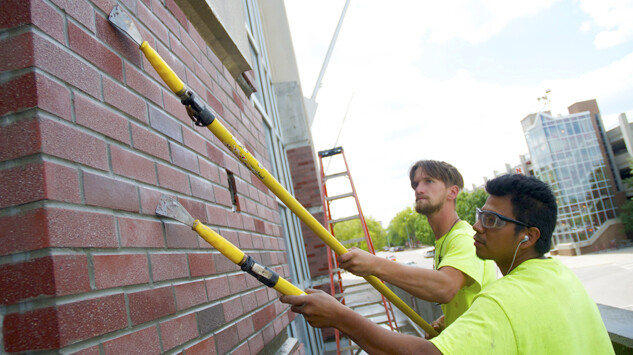 Juan Martinez (right) and Matthew Drazic, both of Masonry Precast Specialty Services, scrape mortar from the face of bricks recently installed on the lower level of UNL's West Memorial Stadium. The duo was using a chemical that softens the mortar for removal.