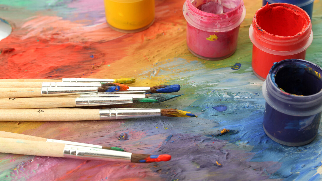 Nine wooden paintbrushes next to multiple colors of paint in cups on a multicolored surface