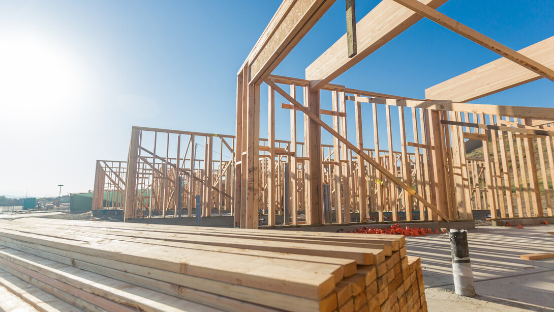 A stack of boards stands in front of the wood frames of three houses on a sunny day.