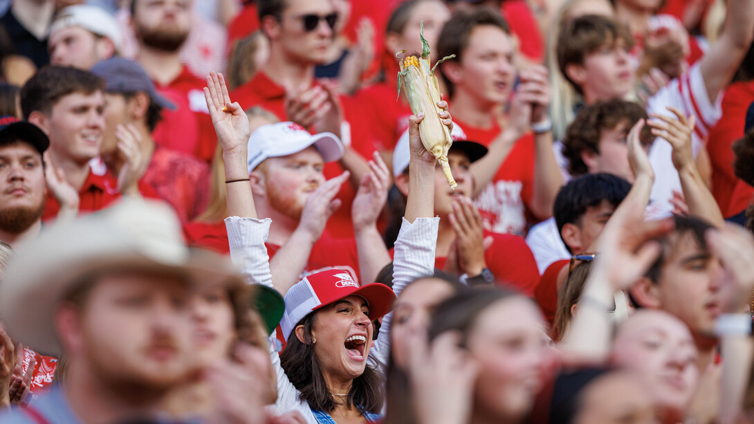 A woman holds her arms in the air, holding an ear of corn, amid a crowd of Husker fans.