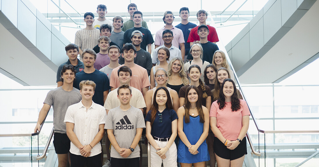 About 30 Husker students — all members of the fifth class of the Investors With Purpose program — stand on a staircase in Howard L. Hawks Hall.