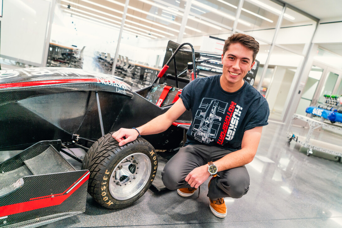 Nick Gauger, a senior mechanical engineering major, wears a Husker Motorsports T-shirt as he poses next to a black-and-red race car in the lower level of Kiewit Hall.