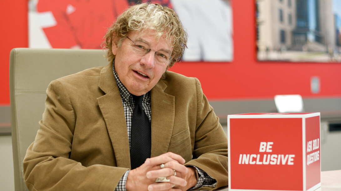 Musician, sound engineer and University of Nebraska–Lincoln alumnus J. E. Van Horne Jr. sits at a table. He is wearing a black-and-white plaid dress shirt, black tie, brown jacket and glasses.