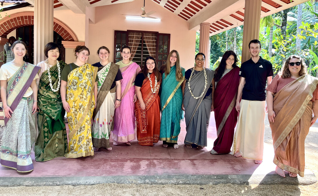 Ten young women and a young man stand outside in traditional Indian clothing.