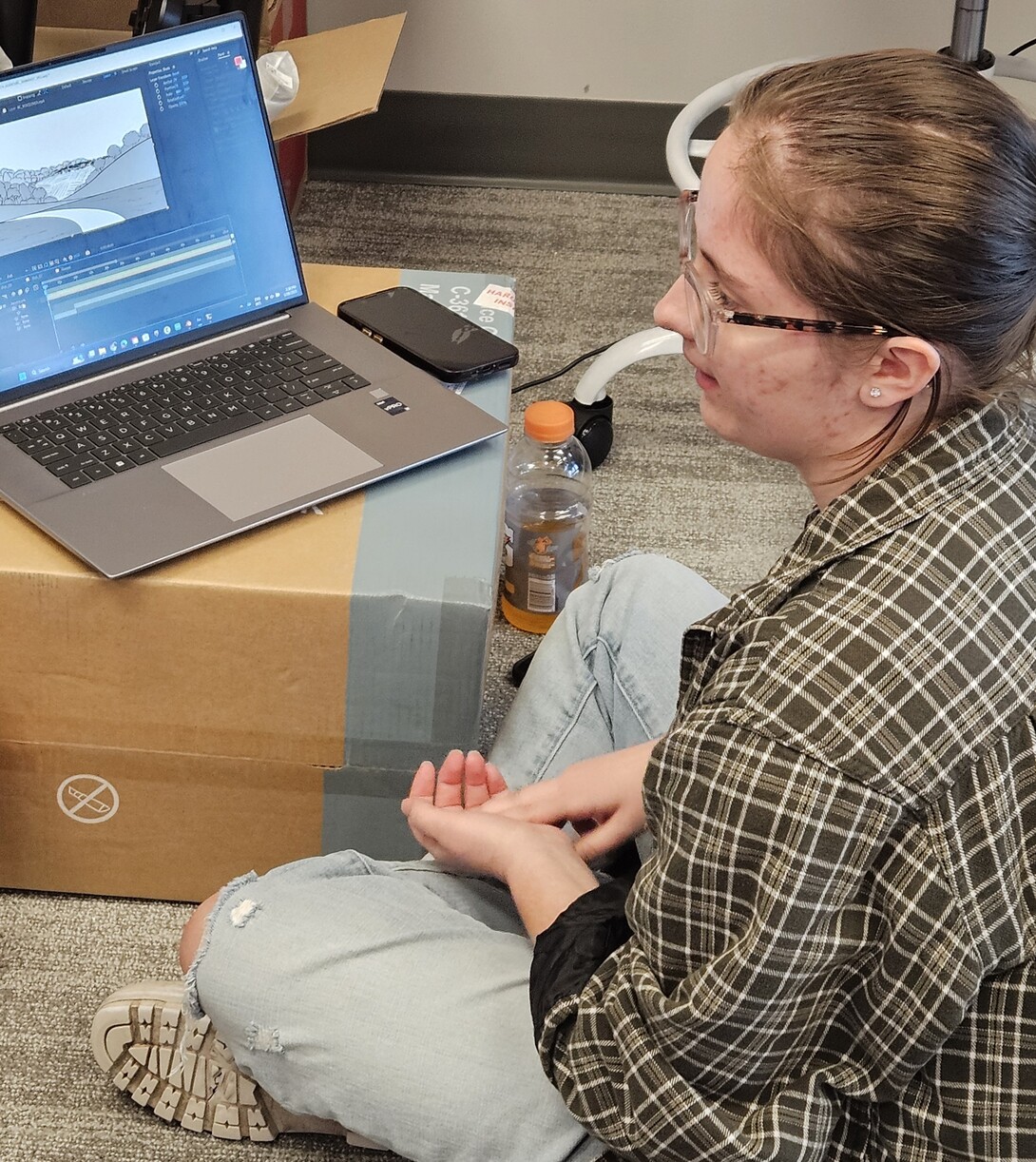 A student in a brown plaid shirt sits on the floor before her laptop, propped on a box.