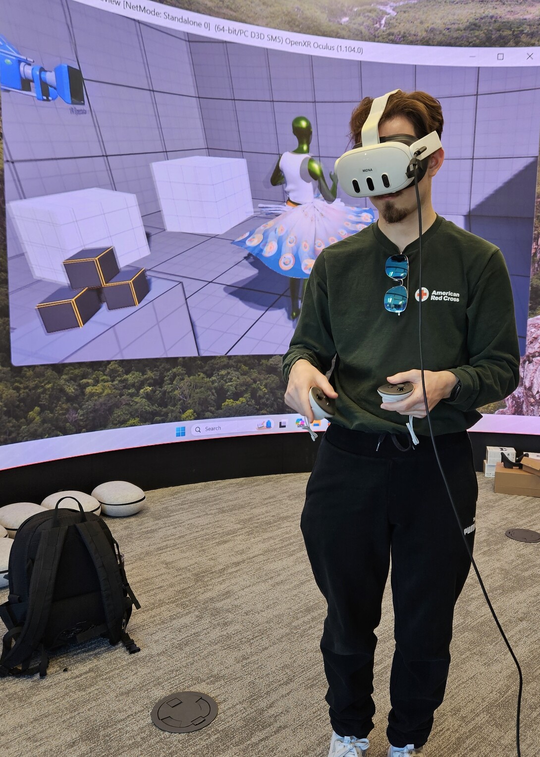 A student in a green shirt and black sweatpants operates a VR headset, standing in front of a screen depicting a 3-D animated dress.
