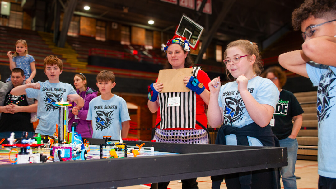 Four members of the Sparks Sharks, a FIRST Lego League robotics team from Wilbur-Clatonia Public Schools, gather around a table in the Coliseum to watch their colorful Lego robot in action. A female judge and a few other people watch, as well.