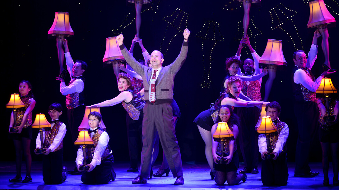 A middle-aged man in a sweater is surrounded by people wearing sparkly vests and bowties, carrying leg lamps, in this scene from "A Christmas Story: The Musical."