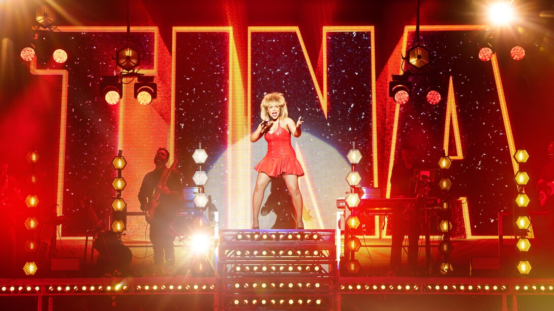 Jayna Elise (as Tina Turner), wearing a red dress, sings into a microphone in front of a rock band and a giant "Tina" sign in a scene from the musical "Tina: The Tina Turner Musical."