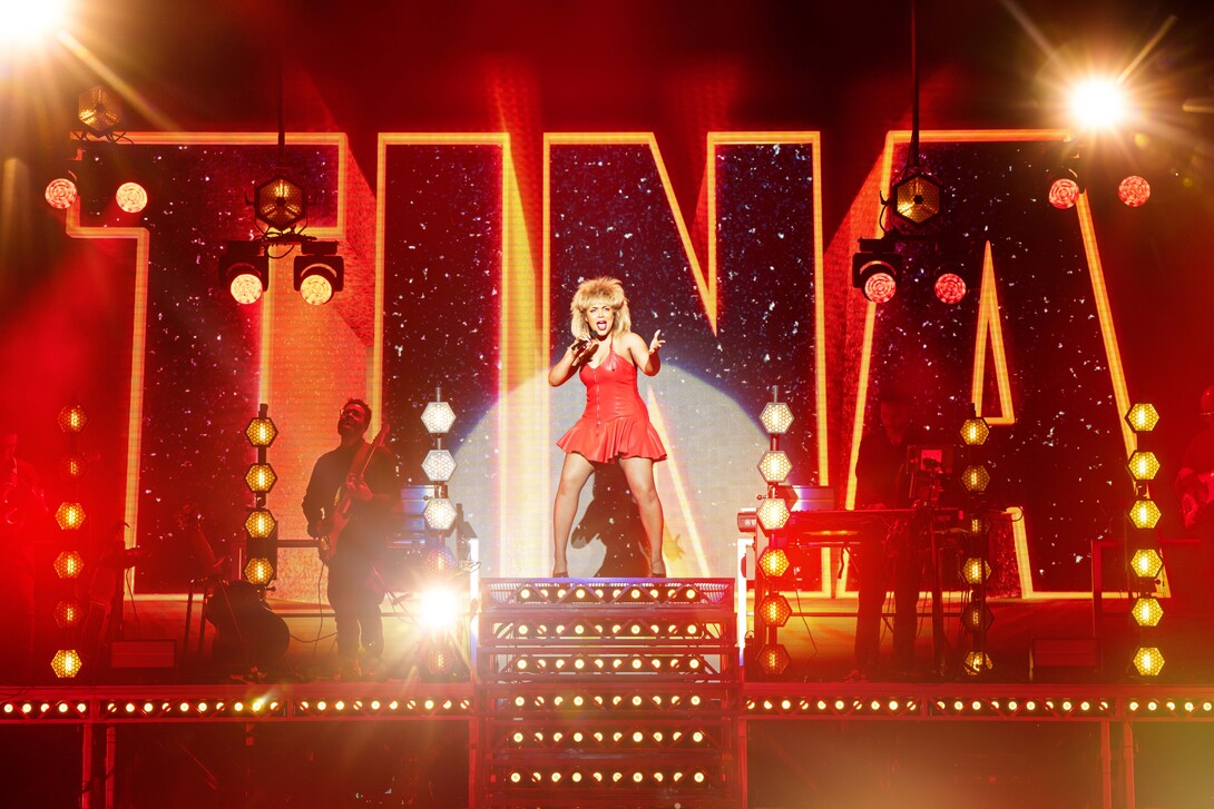 Jayna Elise (as Tina Turner), wearing a red dress, sings into a microphone in front of a rock band and a giant "Tina" sign in a scene from the musical "Tina: The Tina Turner Musical."