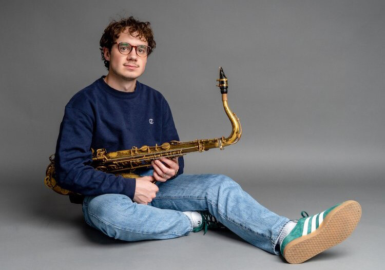 Color portrait of Pete Fucinaro, sitting on the ground with a saxophone.