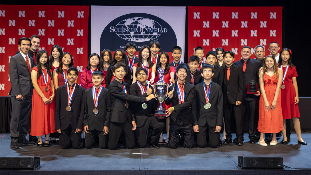 About two dozen students and their coaches from Sierra Vista Middle School in Irvine, California, wearing formal attire, pose with a trophy in front of a Husker background and the Science Olympiad National Tournament logo.