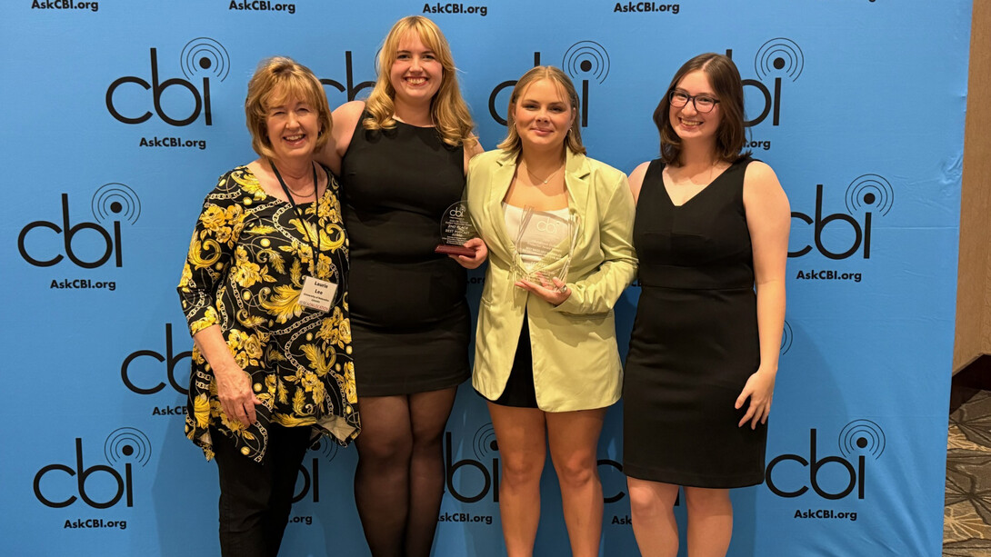 (From left) Nebraska Broadcasting Society faculty adviser Laurie Lee and club members Hayden Hauge, Grace Koch and Katie Mulloy pose with two glass awards in front of a blue background with the words "CBI: AskCBI.org" repeating. They are wearing formal attire.
