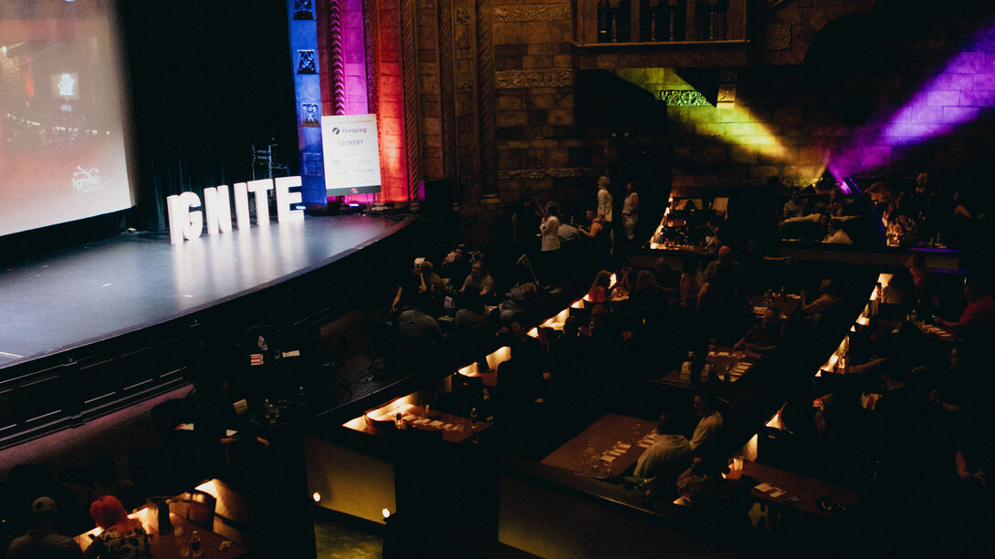 The word "Ignite" graces an empty stage as people wait at tables in the Rococo Theatre.