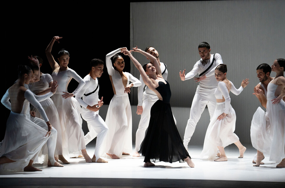 A woman in a black dress dances in front of 11 other dancers, men and women, wearing white.