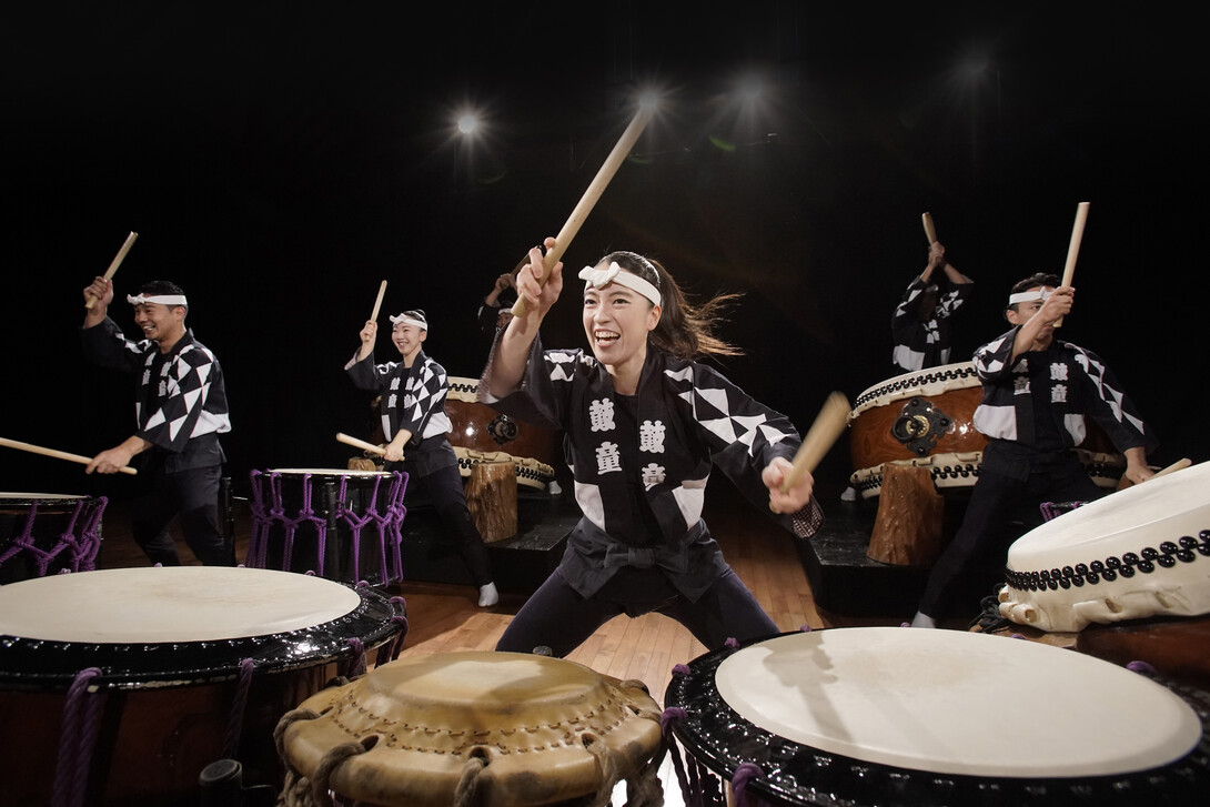 Six drummers dressed in black-and-white outfits with white headbands play ornate taiko drums.