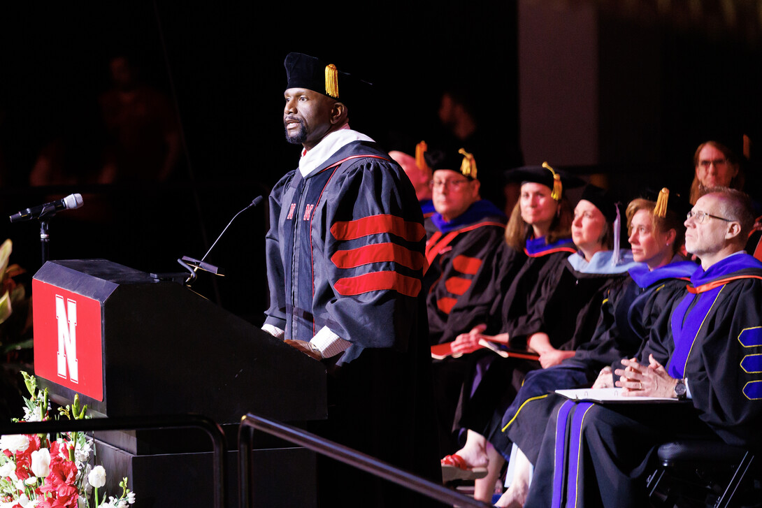DeMoine Adams, wearing graduation regalia, speaks at a lectern as the platform party watches.