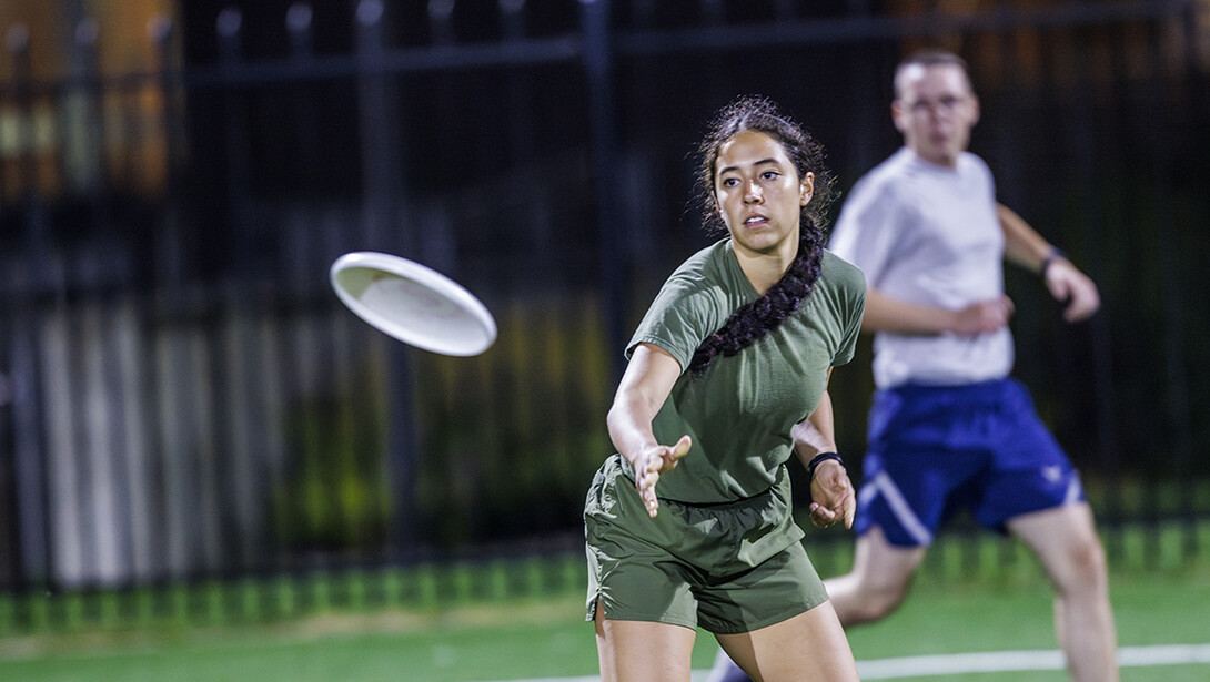 A brunette woman in green athletic wear throws a white frisbee across an indoor field.