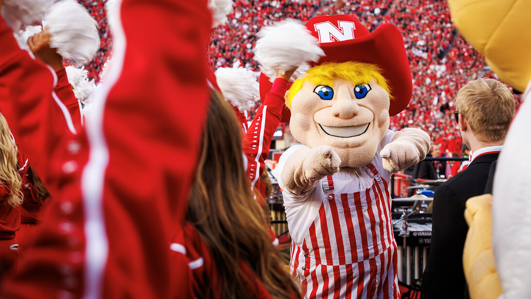 Herbie Husker in red and white striped bib overalls points at the camera during the 2024 Husker football game with Wisconsin.