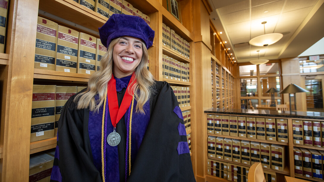 Heather Haratsis, who will graduate from the University of Nebraska College of Law on May 3, stands in the college law library. The background is shelves filled with law books. She is wearing her commencement regalia, which includes a black and purple robe, purple tam (graduation hat) and a medal around her neck with a red ribbon.
