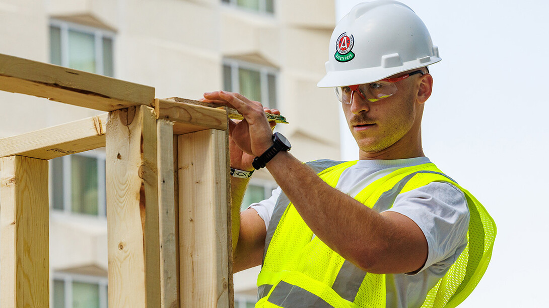 A white man in a light grey t-shirt, green reflective construction vest, white hard had and clear protective glasses writes on 2-by-fours being nailed together.