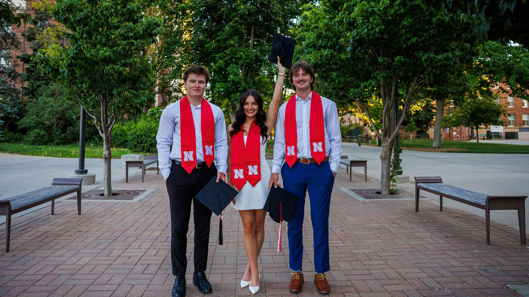 Two men stand around a brunette woman, all in white, outside.