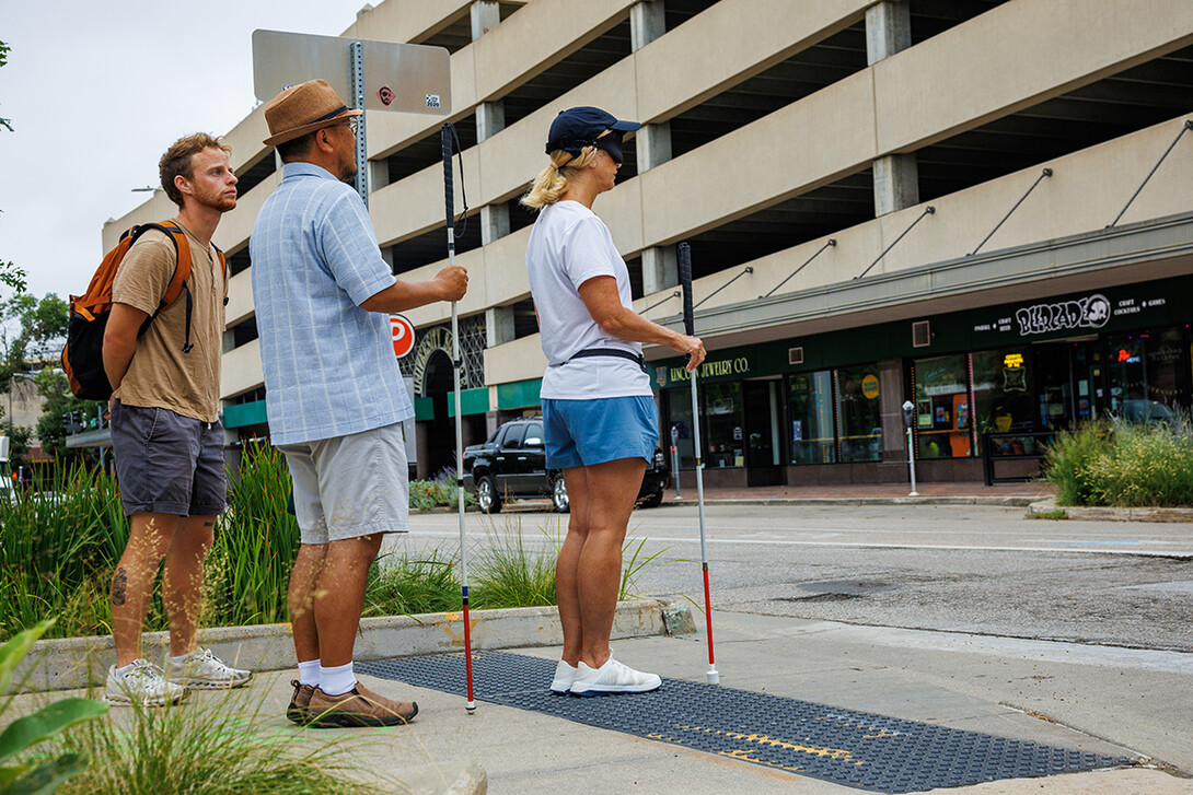 Lori Maestas stands on tactile paving, a textured surface that helps pedestrians with visual impairments know a crosswalk is near. Maestas is working on her posture and where to hold the cane. Victory Klafter (left) and Ko Inamura stand behind Maestas.