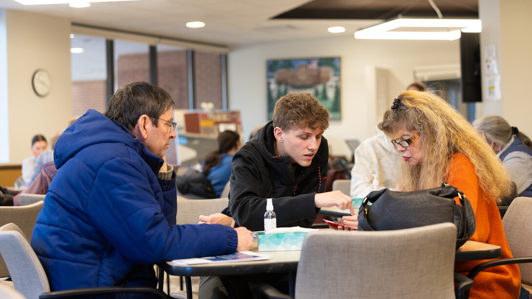 Three people sit around a table as one points to something on a cell phone.