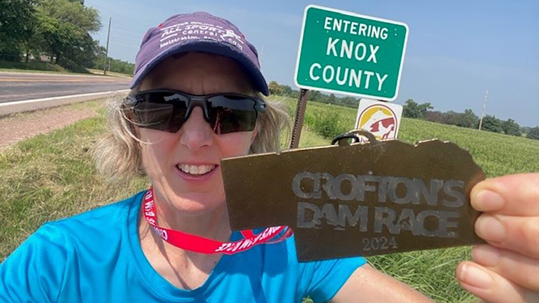 Beth Barry in a blue shirt and purple hat, posing with a medal she recieved after racing Crofton's Dam Race next to the Knox County sign.