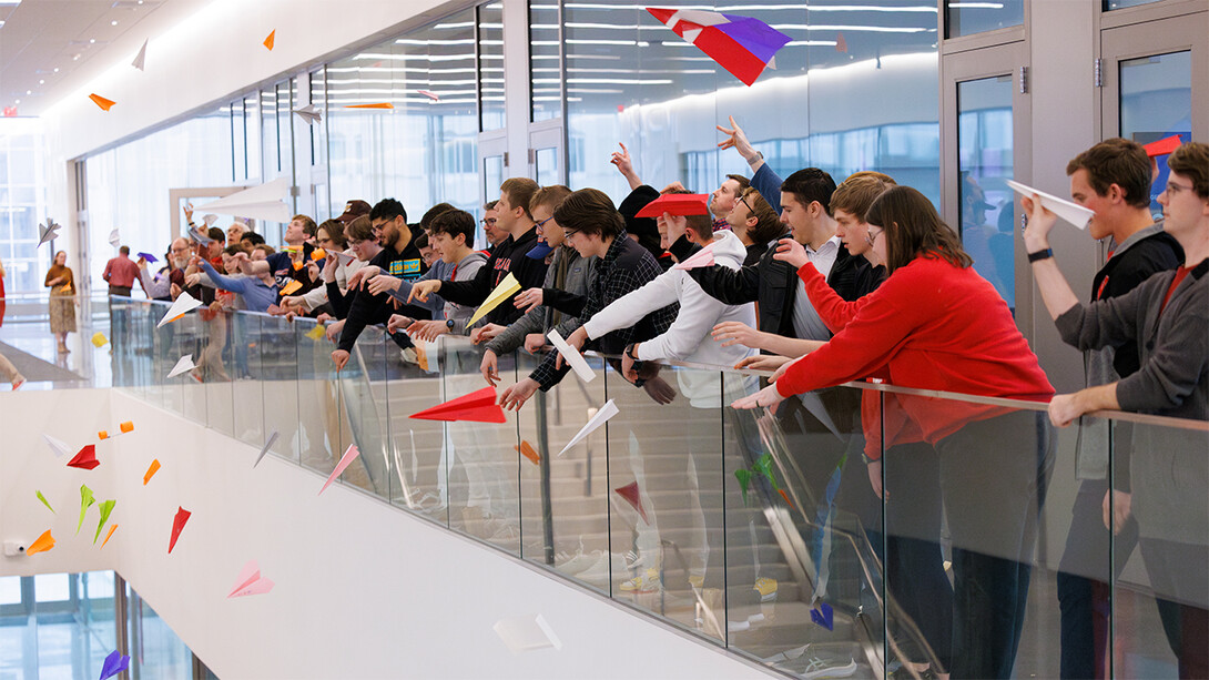 Standing in the open corridor of an upper level, Nebraska Engineering students, staff and faculty toss paper airplanes across the atrium of Kiewit Hall