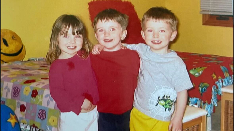 Three children, one girl and two boys, all brunette, pose for a picture together in their childhood bedroom.