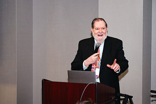 James Gentry, a white man with dark brown hair and a white beard, stands in a black suit while speaking at a podium into a microphone.