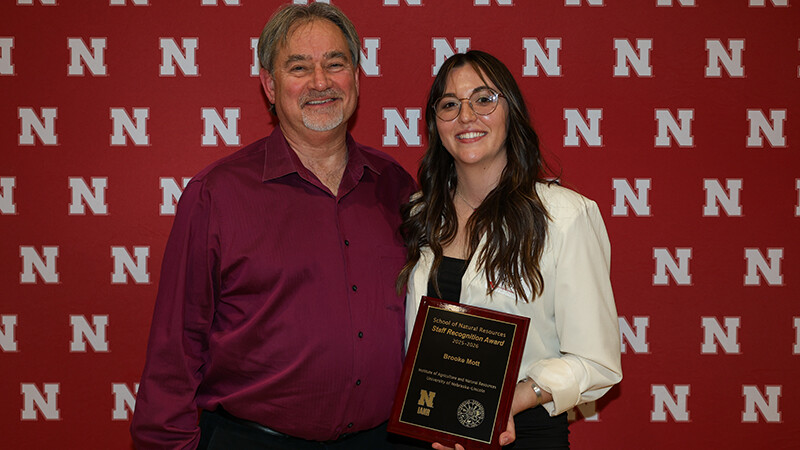 Mark Burbach, a white man with grey hair and a white beard stands in a red button up shirt to the left of Brooke Mott, a white woman with long dark hair and glasses holding a square plaque. 