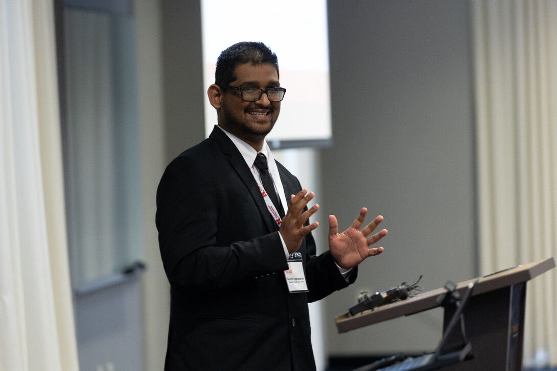 Revanth Raghupatruni, a brown man with black glasses, short black hair and a black moustache, wears a black suit while speaking at a podium.