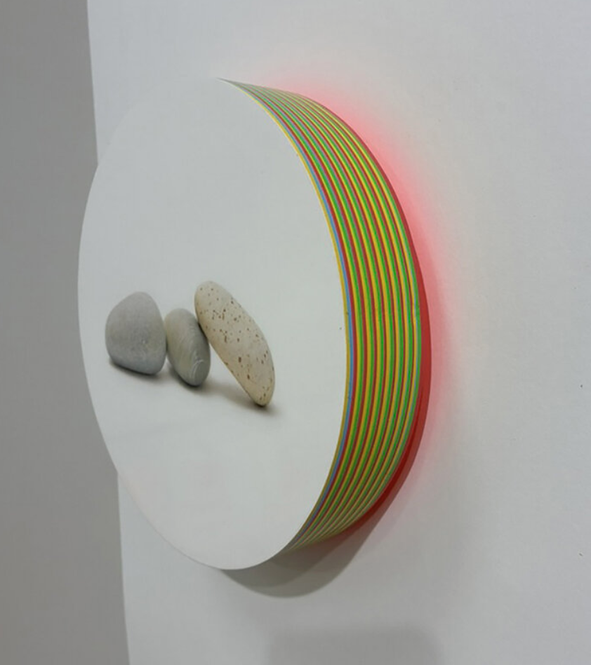 Panel from Francisco Souto's "Eight Million Broken Dreams" art piece. Panel is round showing three rocks sitting on the ground. The base of the piece — which protrudes from the wall — is brightly colored yellow and orange.