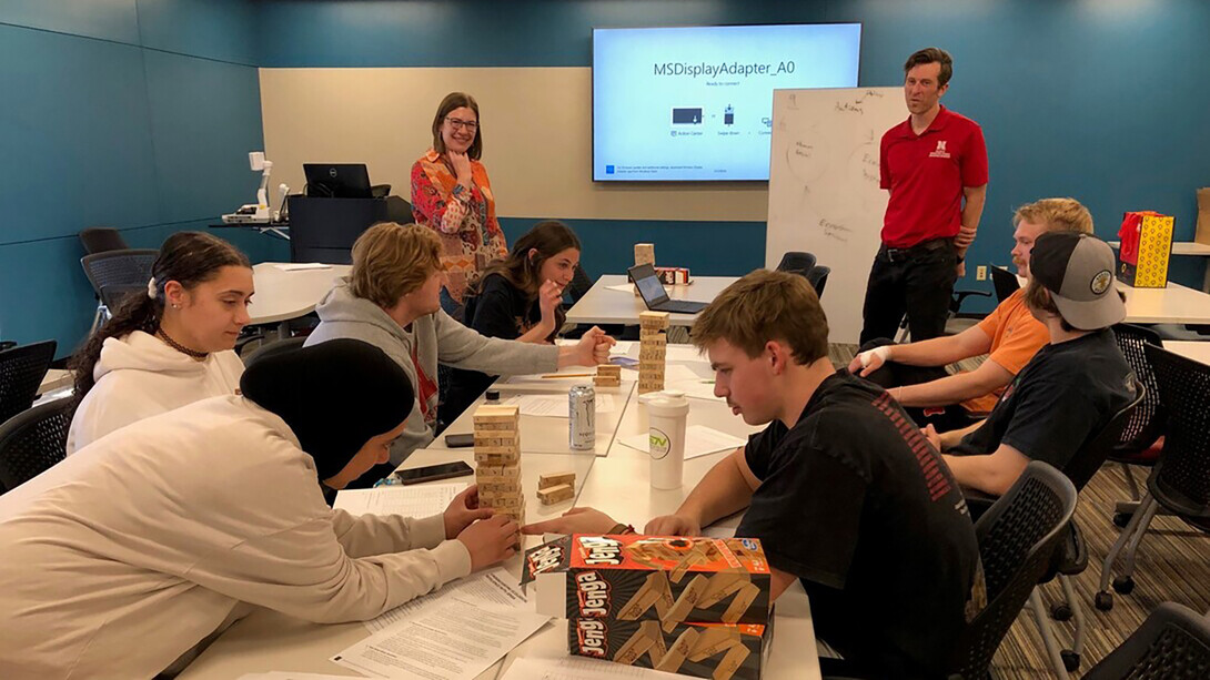 A class of various high school students play different games of Jenga while a woman in a quilted blazer and glasses and a man in a red Nebraska polo supervise.