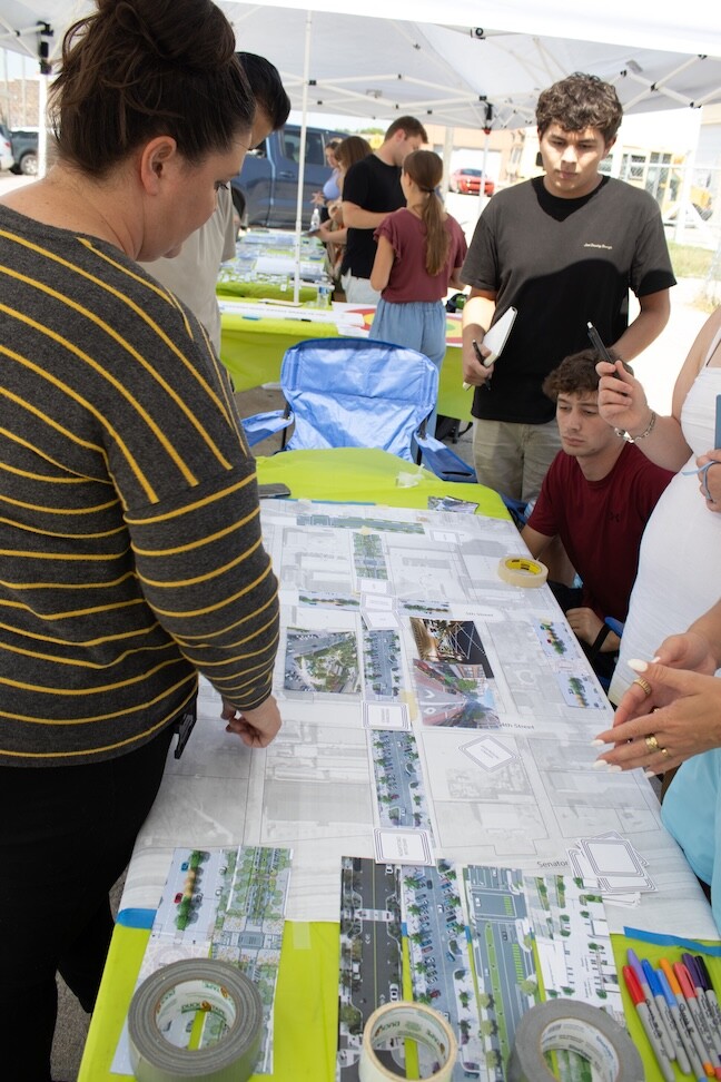 A brunette woman skims informational plans printed out and laid out on a table in front of architecture students.
