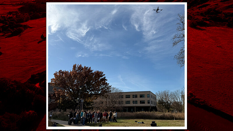 A class of people watch a drone fly in a blue sky.