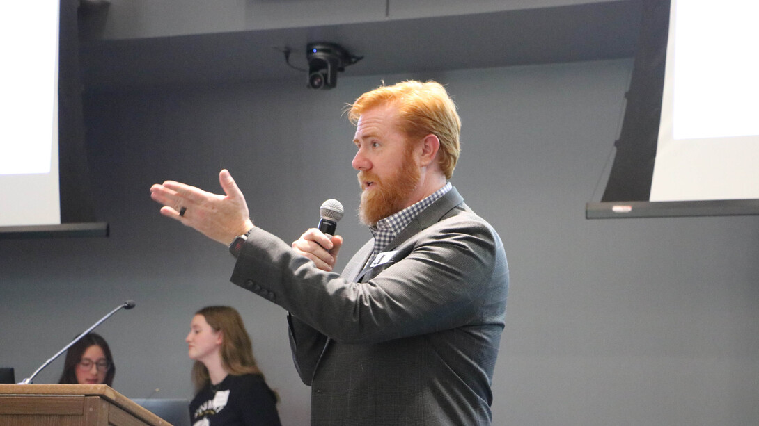 A red-headed man in a grey suit gestures while speaking into a microphone.
