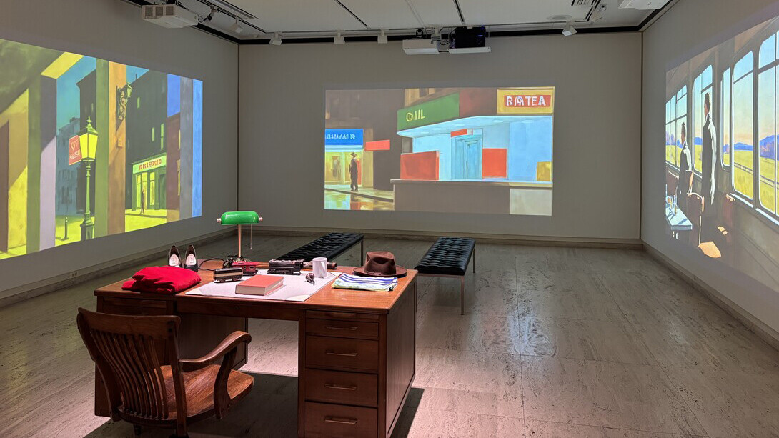 A desk sits in the middle of the Infinite Hopper: An Algorithmic Journey Through Light and Space exhibition in Sheldon Museum of Art. Hopper artworks are projected on three walls.