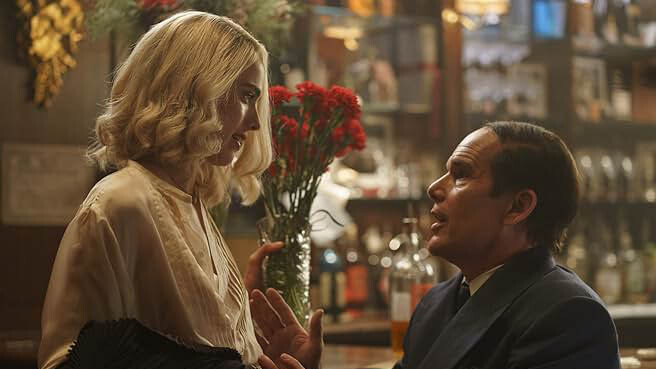 A white, blonde woman in a white dress looks down at a white man with a thin comb-over and a black suit, sitting on a barstool.