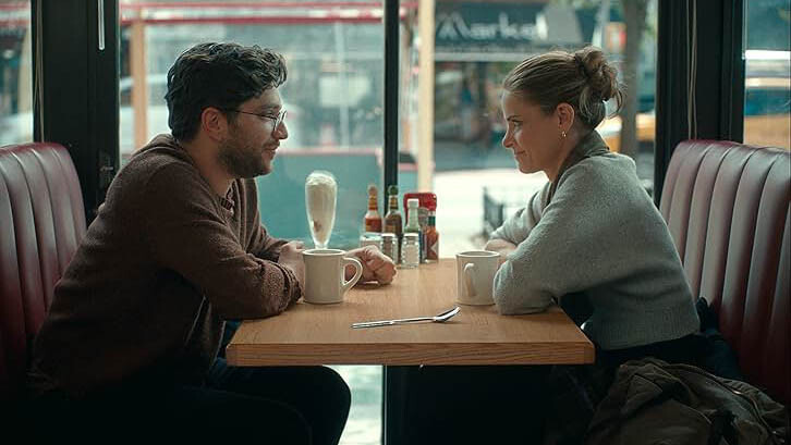 A white man with curly brown hair and a brown bear sits in glasses and a brown sweater in a diner diner booth, across from a white woman with brown hair in a bun, sittingin a light blue sweater.