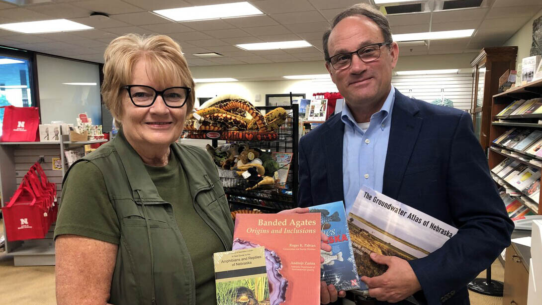 A white woman with short blonde hair and dark glasses poses with a white man with short brown hair, in a navy button up and blazer, both holding multiple small books.