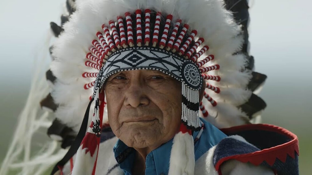 A Native American man in traditional headdress looks at the camera.